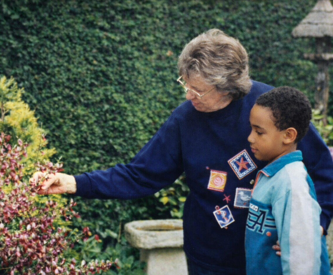 Childhood photo of Alexander J. Lynn, a UK-trained garden designer now based in North Carolina. Restored photograph of Alexander J. Lynn, a UK-trained garden designer now based in North Carolina, as a child. He stands in a garden with his grandmother, her arm extended and her hand touching a native plant.